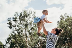 woman lifting her toddley son up into the sky in front of a treeline